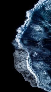 Vertical aerial drone photography of a deep blue ocean wave crashing onto a pitch-black beach, high contrast white foam patterns, moody dark nature aesthetic, minimalist composition for AMOLED screens 4K wallpaper