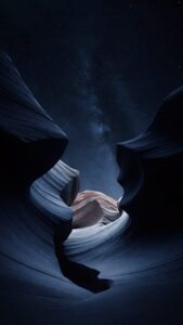 Vertical night landscape photography, looking up through a smooth sweeping slot canyon with dark blue sandstone walls, vast starry night sky and glowing Milky Way galaxy visible through the narrow opening, deep dark aesthetic 4K wallpaper