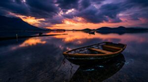 Horizontal 4K digital art of a peaceful sunset over a calm lake, featuring a rustic wooden boat in the foreground, glowing bioluminescent blue water ripples, and dramatic purple and orange clouds reflecting perfectly on the surface, majestic mountain landscape background.