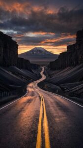 Vertical landscape photography of a long winding asphalt road with yellow double lines leading towards a snow-capped mountain peak at sunset, flanked by dark rocky cliffs, cinematic moody lighting, 4K travel wallpaper.