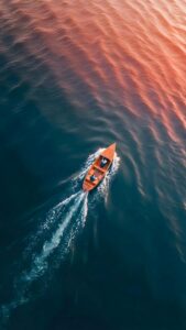 Aerial Boat Ocean Sunset Wallpaper 4K, beautiful top-down drone photography digital art of a small wooden boat sailing in dark blue ocean water with warm orange sunset reflections.