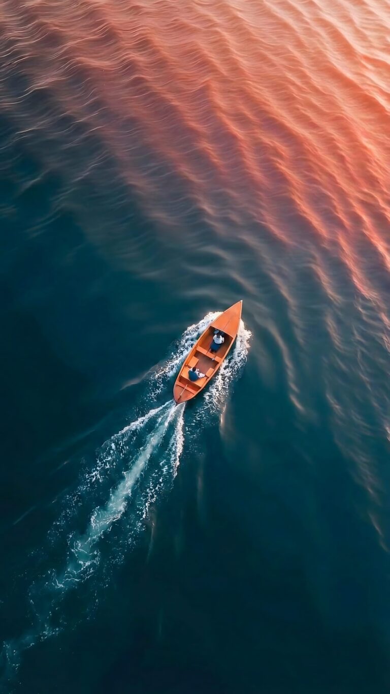 Highly detailed aerial drone photography of a small boat sailing through deep blue ocean water reflecting warm orange and pink sunset colors aesthetic 4K ultra HD mobile wallpaper