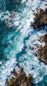 Download free 4K mobile wallpaper featuring an aerial view of turquoise sea waves crashing against rocky shores for a fresh summer look