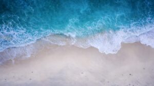 Highly detailed 4K aerial drone photography of beautiful turquoise blue ocean waves crashing on a pristine white sand beach, optimized as a cool desktop wallpaper.