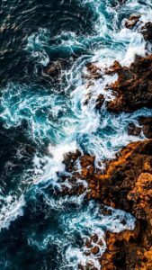 Highly detailed 4K wallpaper featuring an aerial top-down view of deep blue ocean waves crashing against jagged orange coastal rocks, creating bright white foam and a beautiful high-contrast nature scene