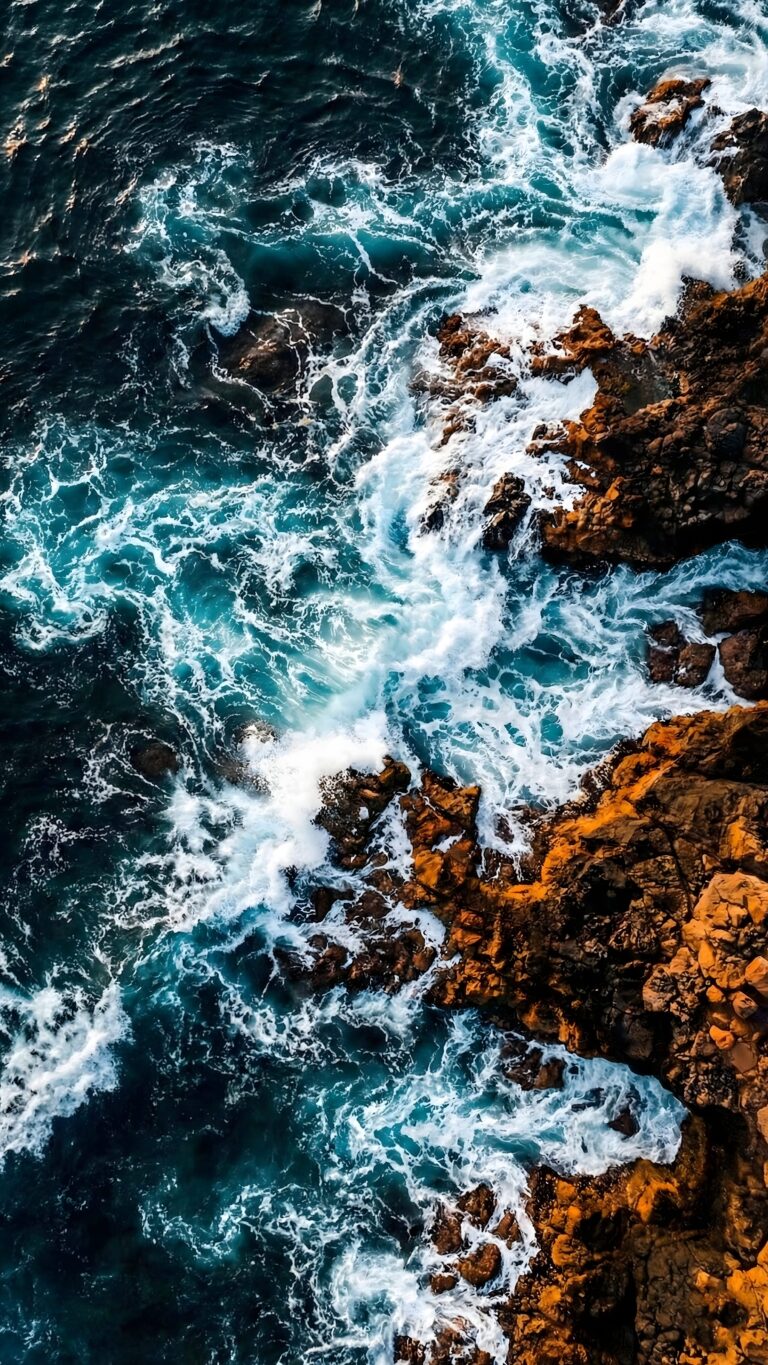 Highly detailed 4K wallpaper featuring an aerial top-down view of deep blue ocean waves crashing against jagged orange coastal rocks, creating bright white foam and a beautiful high-contrast nature scene
