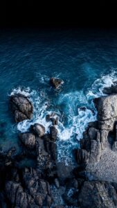 Dark Ocean Waves Aerial Wallpaper 4K, cinematic top-down drone photography digital art of deep blue ocean waves crashing against dark rugged rocks, dark AMOLED background at the top.