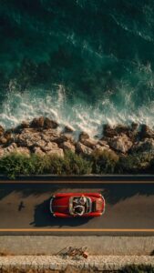 Download free 4K mobile wallpaper of an aerial top-down view featuring a classic red convertible car driving on a coastal road next to beautiful turquoise ocean waves