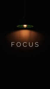 A high-resolution 4K mobile wallpaper featuring the word FOCUS illuminated by a single hanging pendant light against a deep black background, perfect for AMOLED screens.