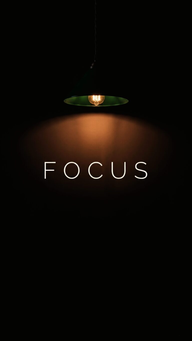A high-resolution 4K mobile wallpaper featuring the word FOCUS illuminated by a single hanging pendant light against a deep black background, perfect for AMOLED screens.