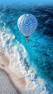 A high-resolution 4K mobile wallpaper of a blue and white patterned hot air balloon flying over clear turquoise ocean waves and a sandy beach.