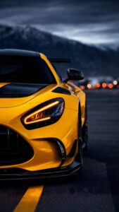 Front close-up of a bright yellow luxury sports car with glowing LED headlights parked on a road with a dramatic mountain background.