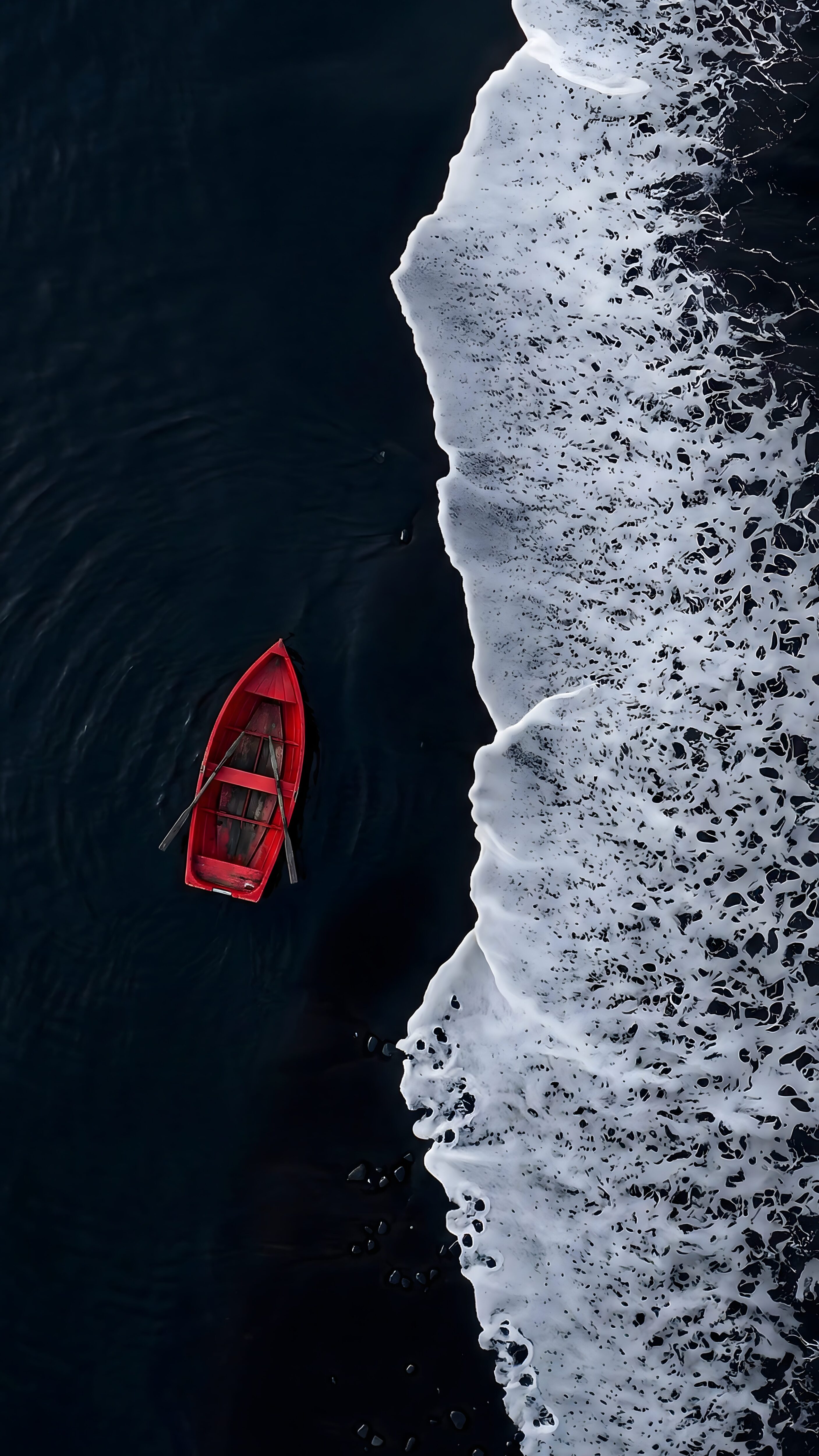 An aerial top-down view of a small bright red rowboat floating on extremely dark water right next to crashing white foamy ocean waves.