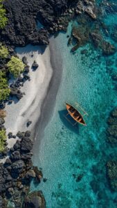 A high-resolution 4K aerial mobile wallpaper of a traditional wooden boat floating in crystal clear turquoise water next to a dark sandy and rocky coastline.