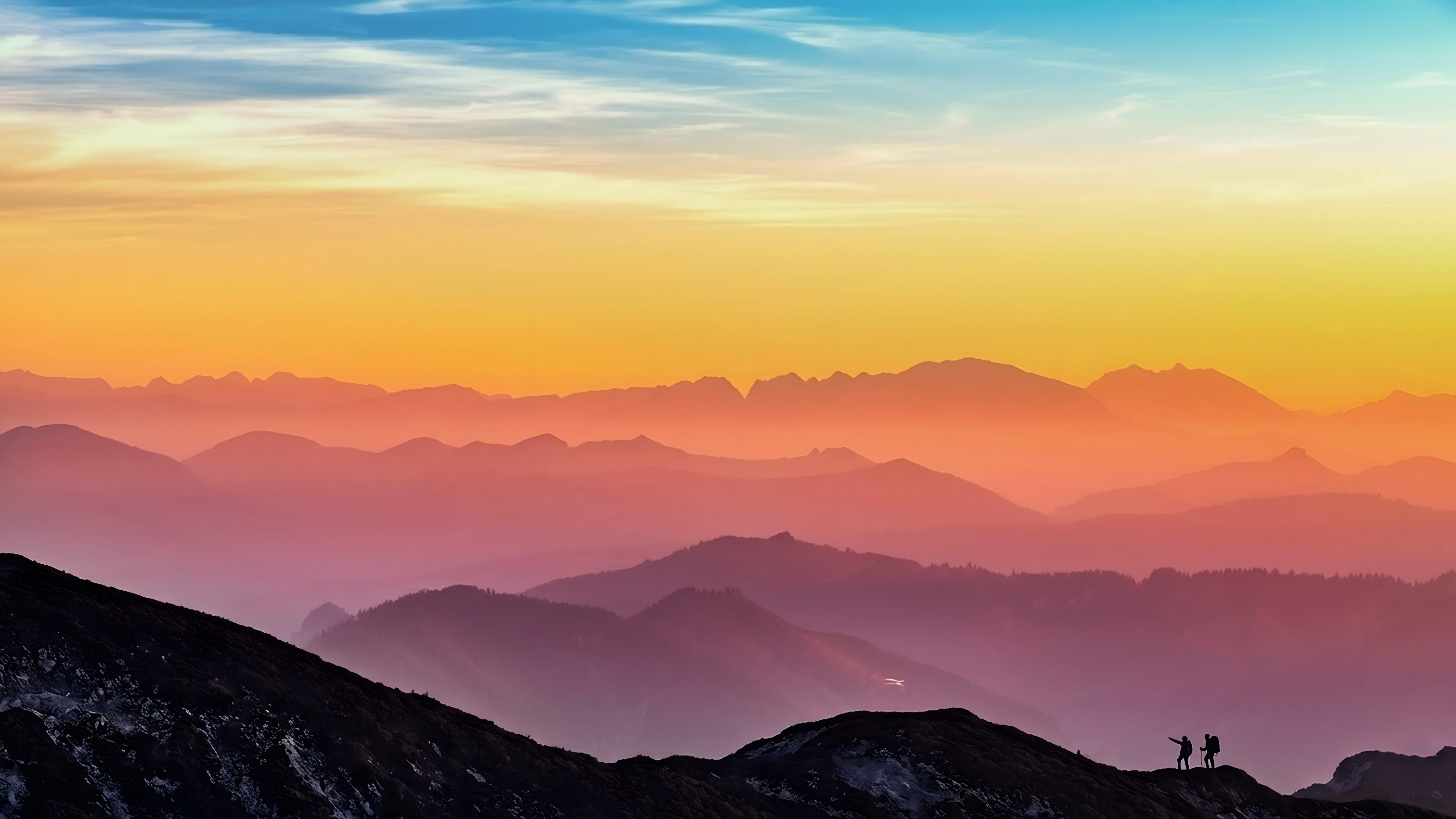 Vibrant 4K desktop wallpaper of colorful misty mountains during sunset with silhouettes of two hikers on a ridge.