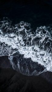 Top-down 4K mobile wallpaper of white sea foam crashing onto a black sand beach with a dark ocean aesthetic.