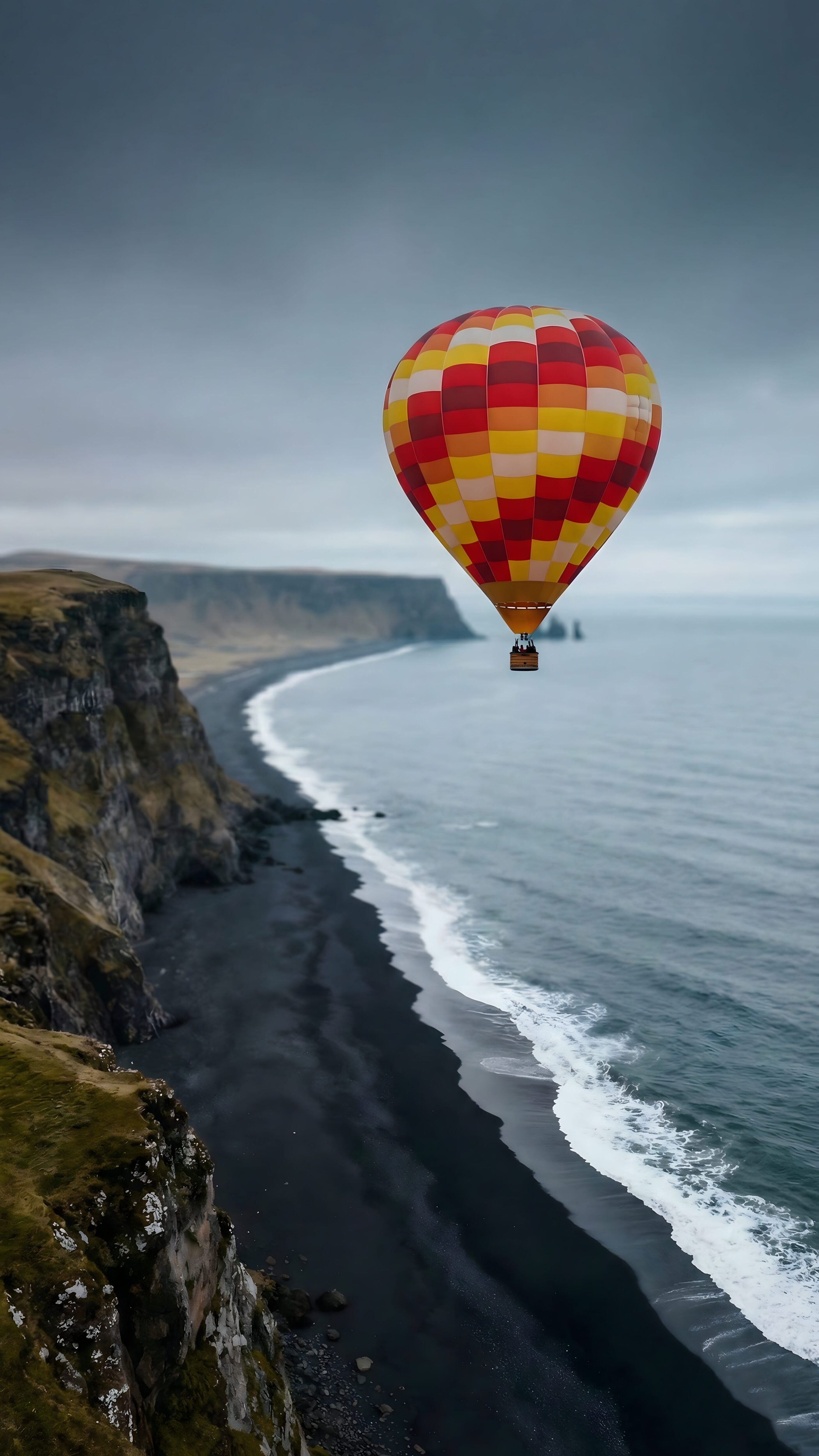 4K mobile wallpaper of a colorful hot air balloon flying over a dramatic black sand beach, steep green cliffs, and ocean under a moody sky.