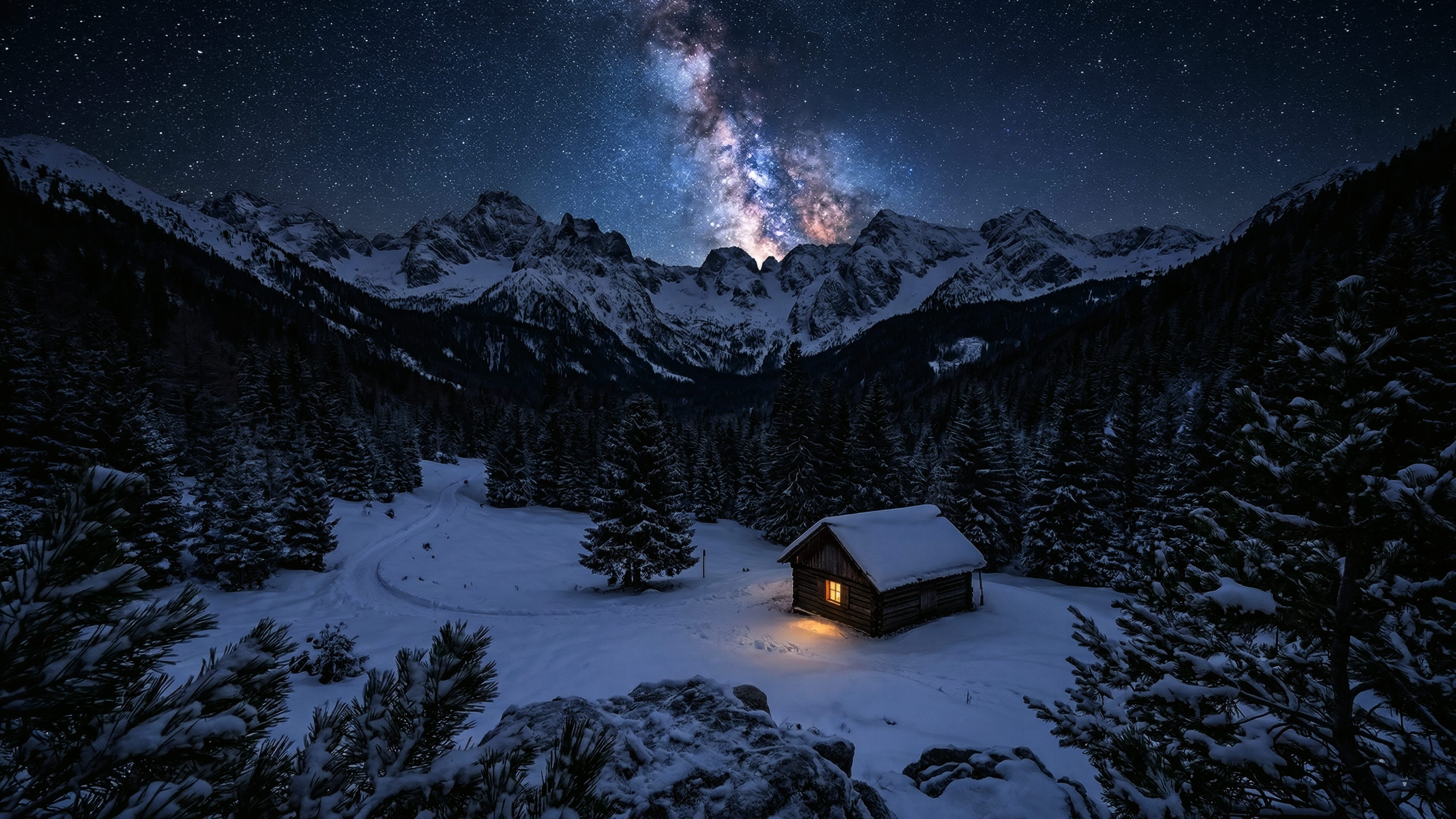 A high-resolution desktop wallpaper featuring the Milky Way galaxy stretching over snow-covered mountains and a small wooden cabin with a warm glowing light.