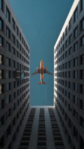 Ultra-clear 4K mobile wallpaper showing an orange airplane flying directly between two symmetrical high-rise city buildings under a blue sky.