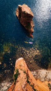 A high-resolution 4K mobile wallpaper featuring a top-down view of red rock cliffs, a sea stack, and clear blue water with a yellow kayak.