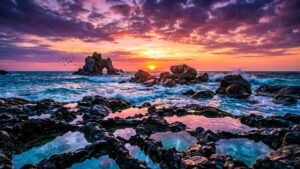 A wide 4K desktop wallpaper of a rocky coastline at sunset, featuring crashing waves, a rock arch, and foreground tide pools reflecting a purple and orange sky.