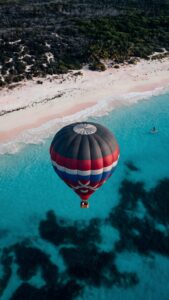 Aerial view of a colorful hot air balloon flying over turquoise ocean water and a white sand beach with tropical trees.