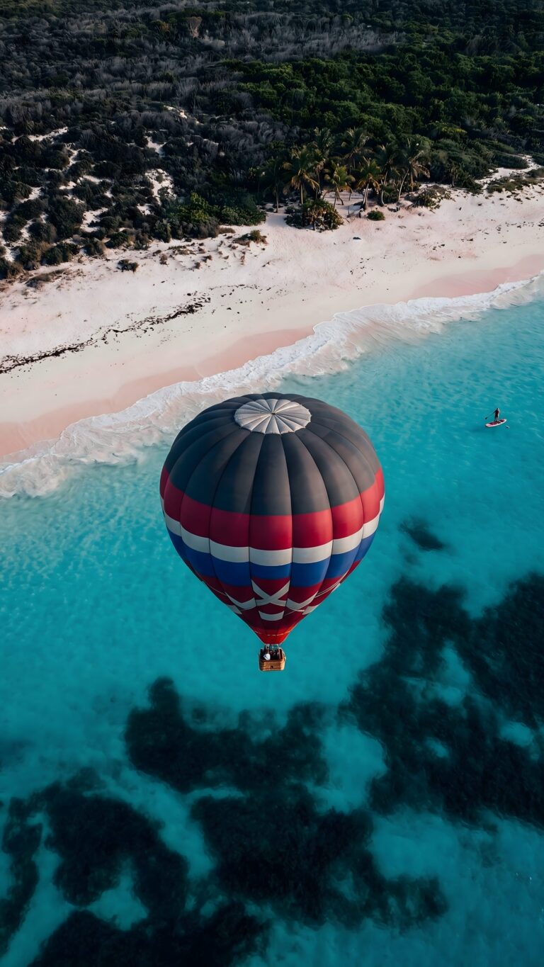 Aerial view of a colorful hot air balloon flying over turquoise ocean water and a white sand beach with tropical trees.