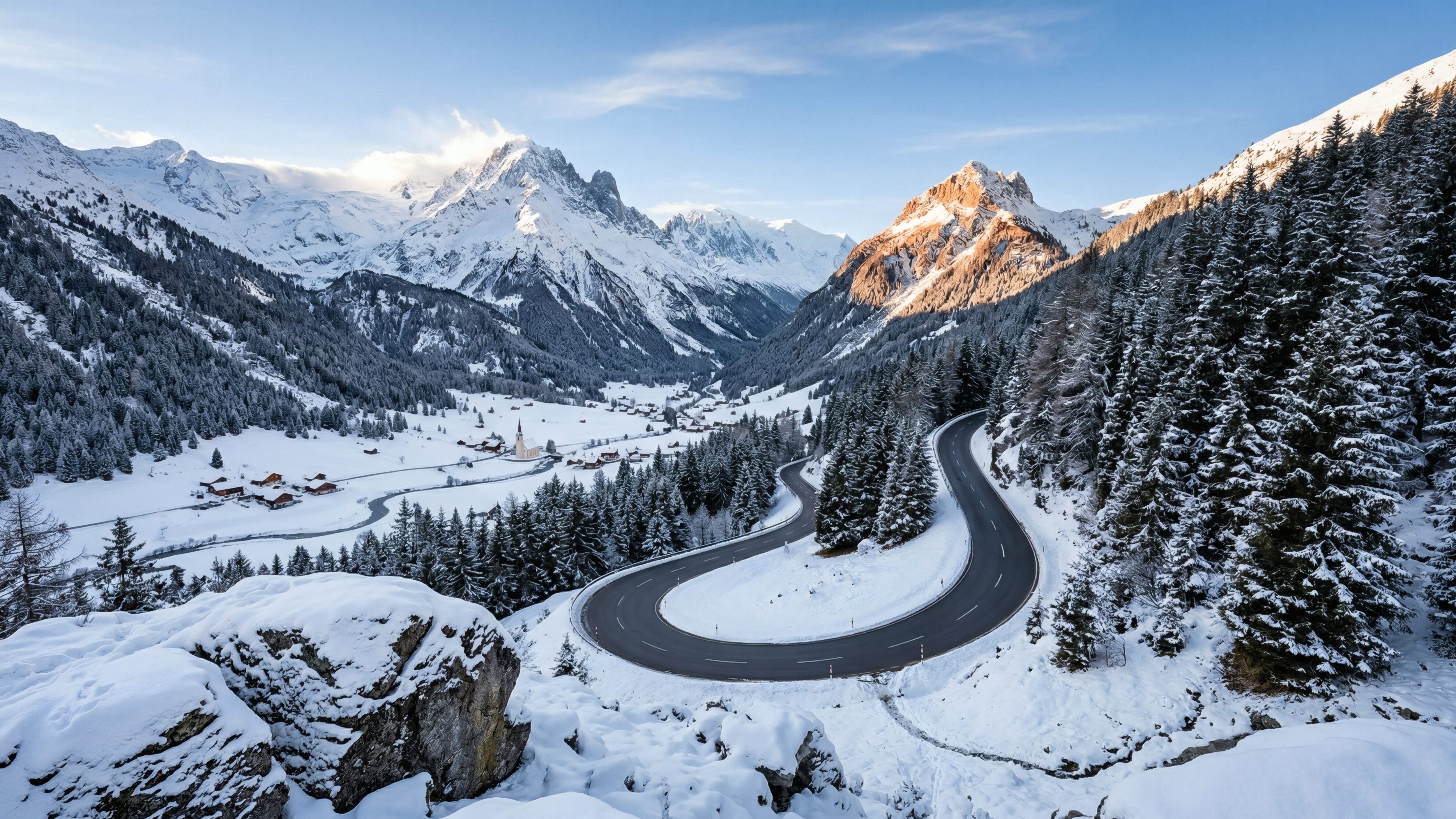 A high-resolution 4K desktop wallpaper featuring a winding asphalt road through a deep snowy mountain valley with pine forests and sun-lit peaks.