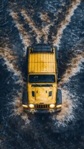 A high-resolution 4K aerial mobile wallpaper of a bright yellow Jeep Wrangler driving through deep river water, creating a muddy wake.