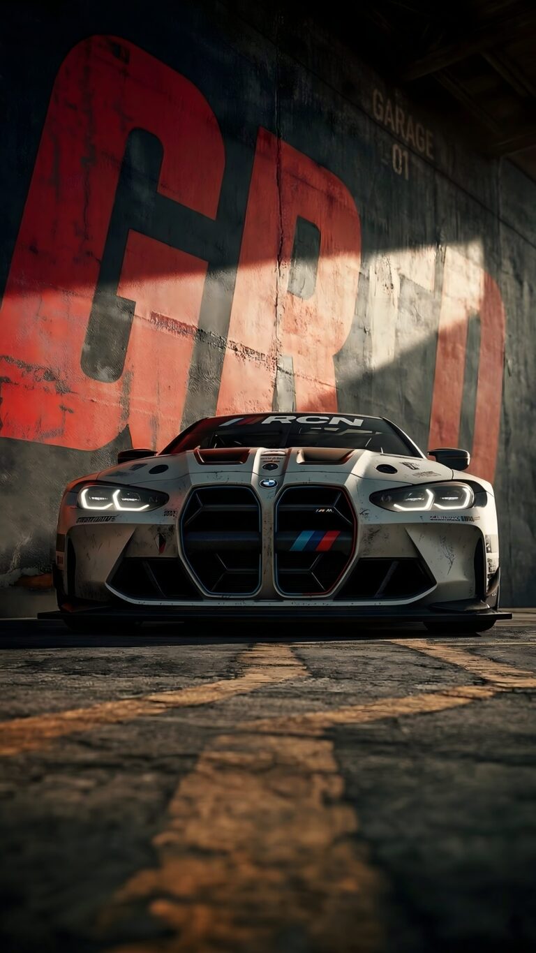 Front view of a dirty, heavily modified white BMW race car with glowing headlights parked in an industrial garage with large red graffiti on the wall.