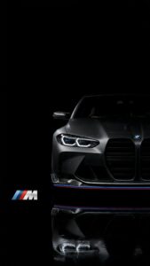 Front view of a sleek matte black BMW M series car with illuminated headlights and an M logo reflection on a pitch-black background.