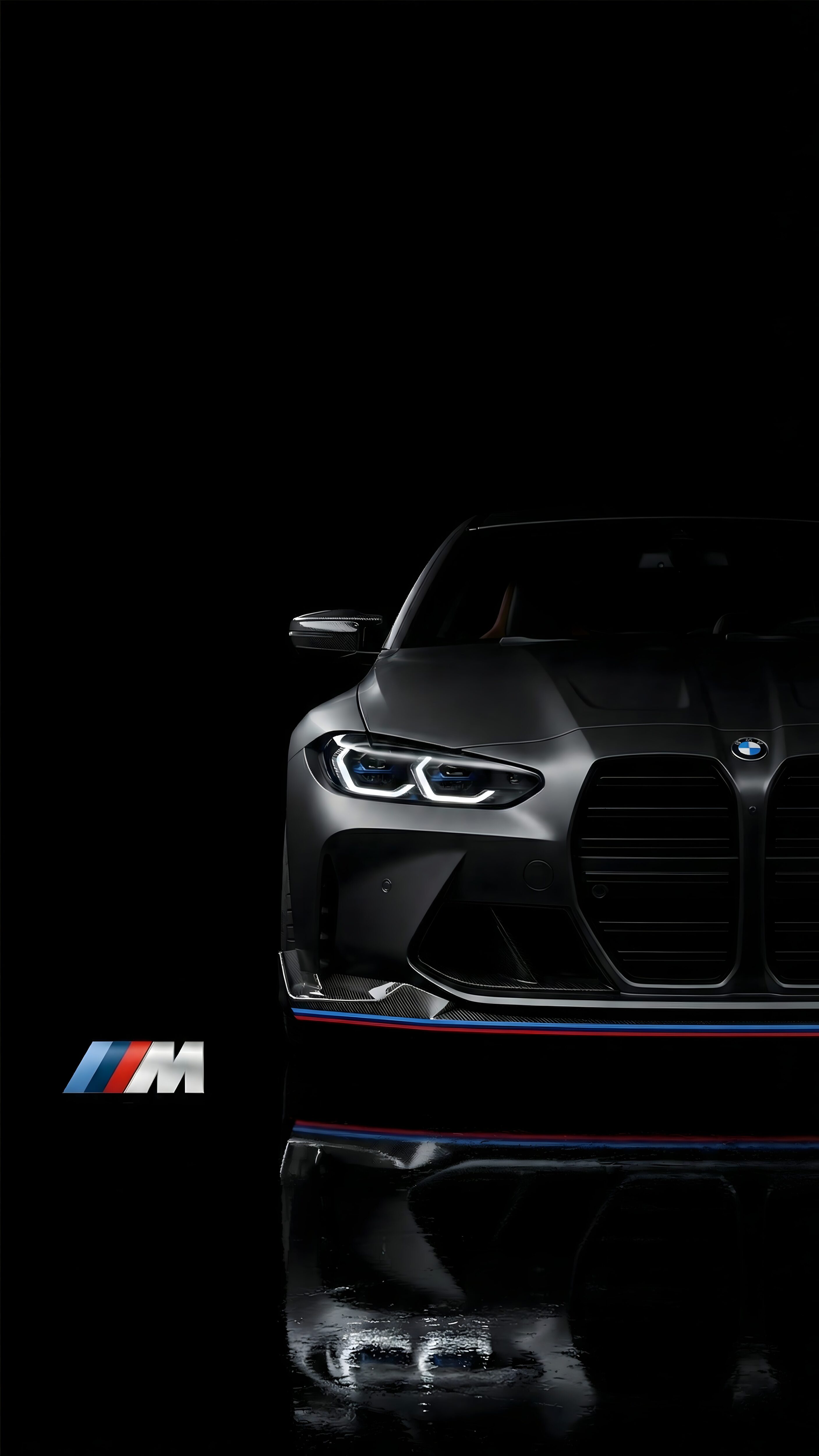 Front view of a sleek matte black BMW M series car with illuminated headlights and an M logo reflection on a pitch-black background.