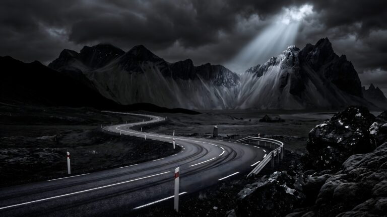 A winding road leading towards sharp, rugged mountain peaks under heavy dark clouds, illuminated by a single bright sunbeam.