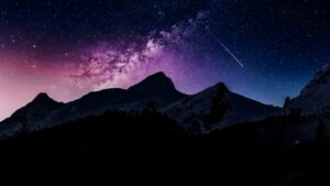Silhouette of mountain peaks against a vibrant purple and blue night sky filled with stars, the Milky Way, and a shooting star.
