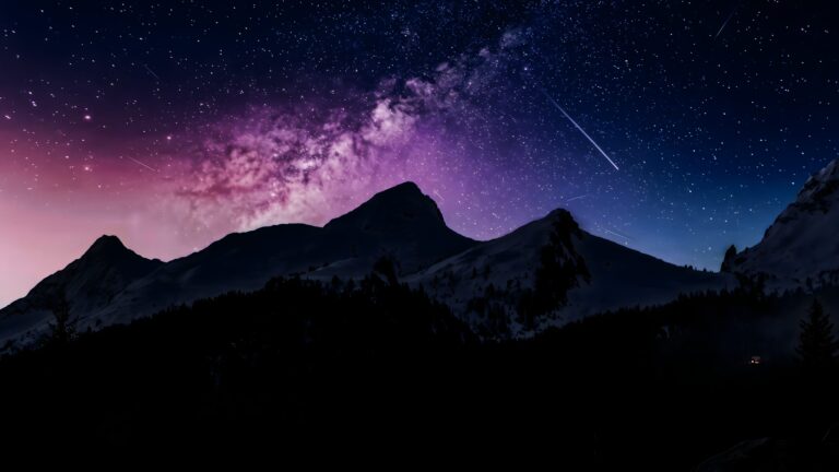 Silhouette of mountain peaks against a vibrant purple and blue night sky filled with stars, the Milky Way, and a shooting star.