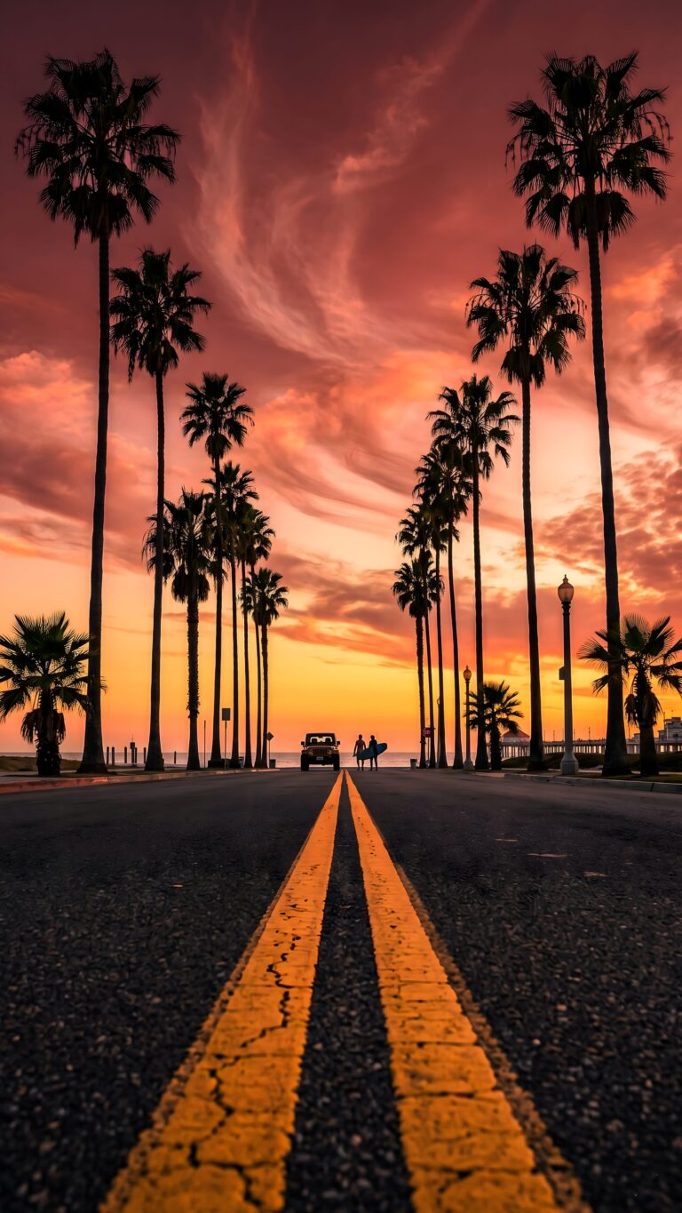 A vertical view down a road lined with tall palm trees leading to a beach, featuring a glowing orange and pink sunset, a parked jeep, and two surfers in silhouette.