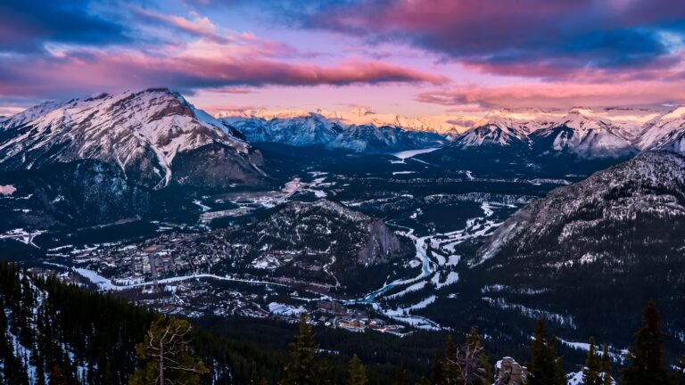 Panoramic view of a snow-capped mountain range at sunset with a winding river and valley town below.