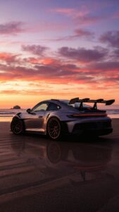 Silver Porsche 911 GT3 RS parked on a wet beach reflecting a vibrant pink and orange sunset sky.