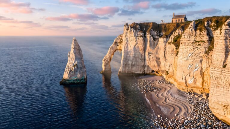 Breathtaking view of white chalk cliffs featuring a natural rock arch, a tall sea stack, and a small cliffside building during a vibrant golden hour sunset.