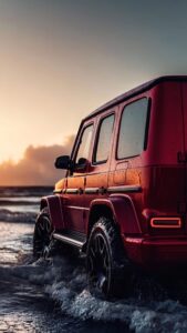 A bright red Mercedes-Benz G-Wagon driving through splashing ocean waves during a vibrant golden hour sunset.