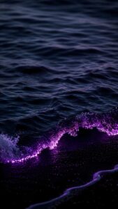 A dark ocean wave crashing onto the beach, glowing with intense bioluminescent neon purple light against the midnight water.