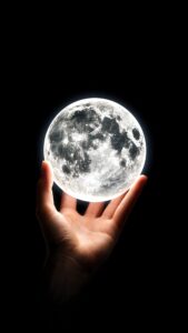 A person's hand glowing under the light of a bright, detailed full moon held in their palm, set against a completely black background.